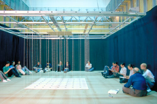 An installation made of parallel threads and squares. All around sit people