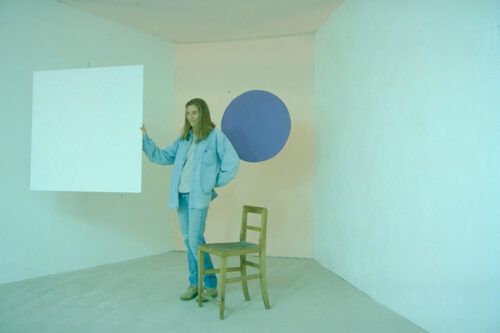 Eine Frau hält ein Quadrat. Neben ihr steht ein Stuhl, hinter ihr ist ein blauer Kreis